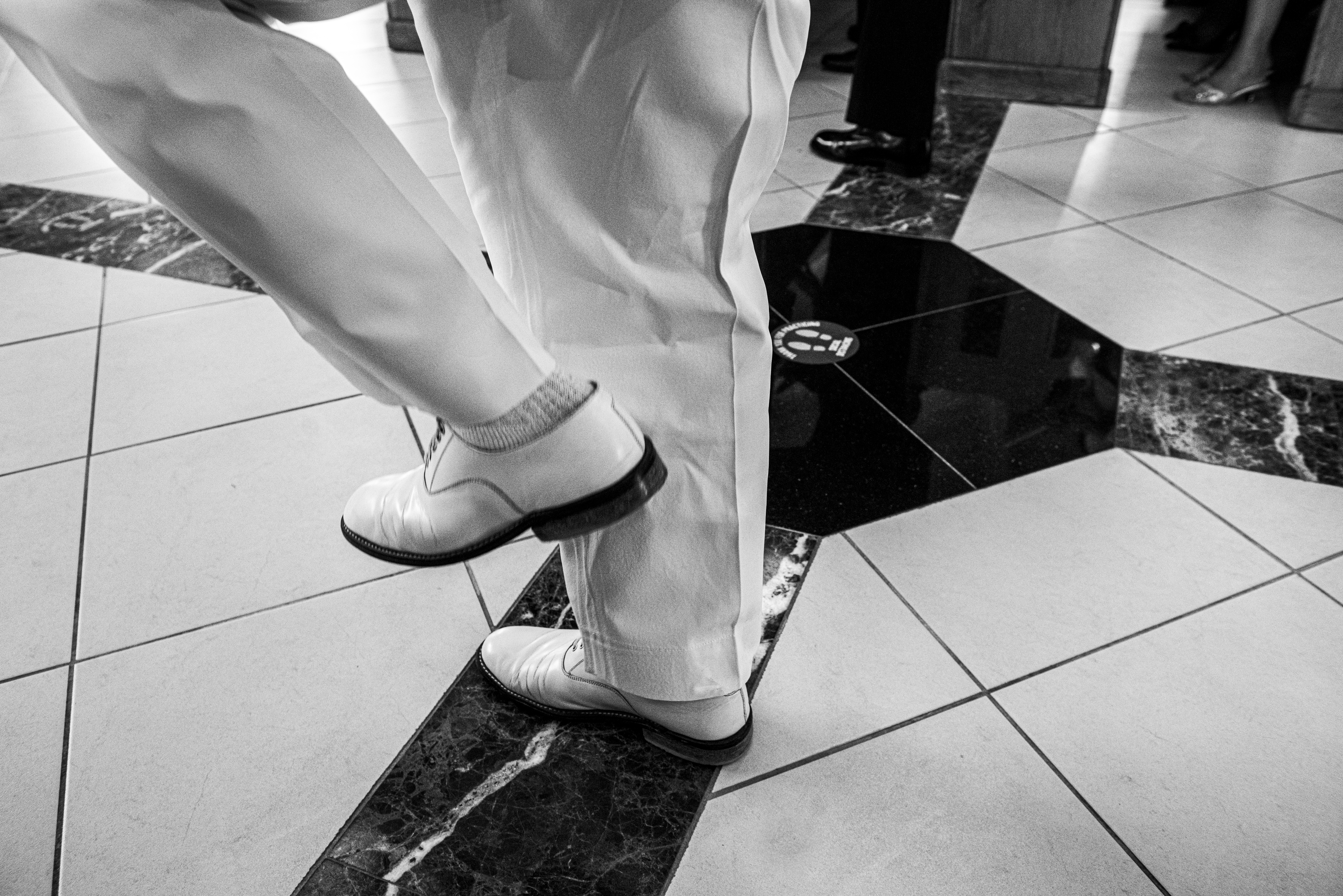 Black and White photo by Hasna Muhammad over white shoes and white pants marching on tiled floor.