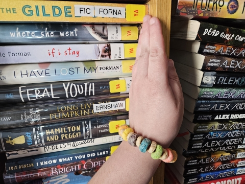 pride month bracelet on a wrist in front of a bookshelf