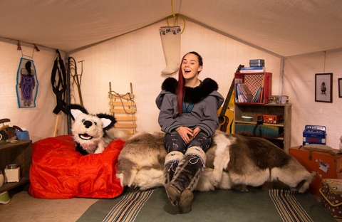 Image of the main set of Anaana's Tent. Host Rita Claire sits in a cloth tent surrounded by books, instruments, and Inuit tools and art. She is accompanied by a large muppet-style dog puppet sitting on a bean bag.