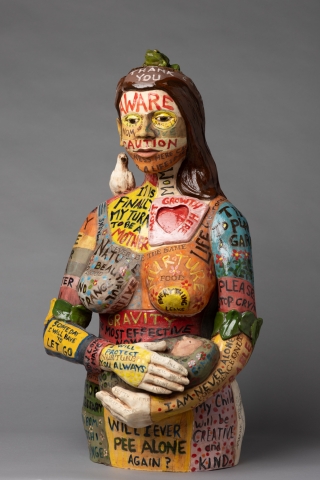 Ceramic sculpture of a female body holding a child with a bird perched on the shoulder and a frog on the head. The body is split into colorful patches with thoughts about motherhood filling each space.